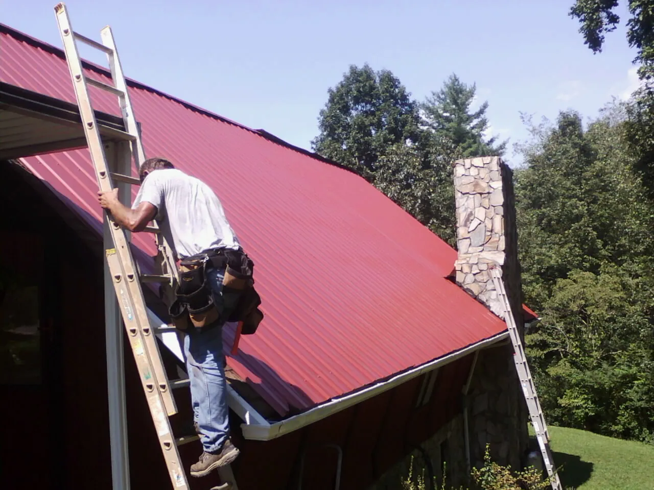 Roofer on a ladder beside a red metal roof and stone chimney — Soffit & Fascia Repair by Roofing Brothers in Dedham